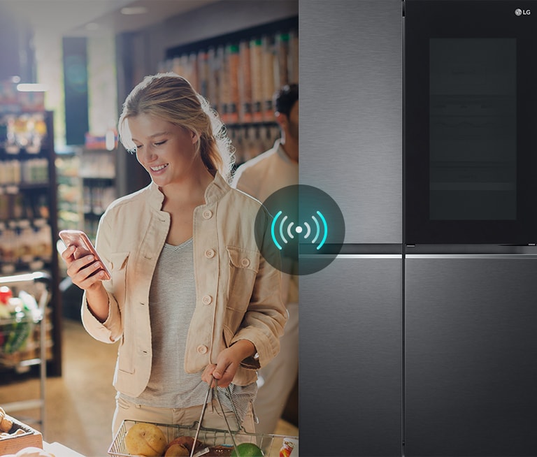 Woman using a phone while shopping next to an LG smart refrigerator that has a Wi-Fi connectivity icon.