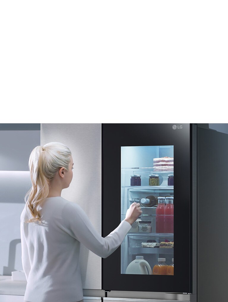 Person interacting with an LG smart refrigerator featuring a transparent door revealing various food items inside.