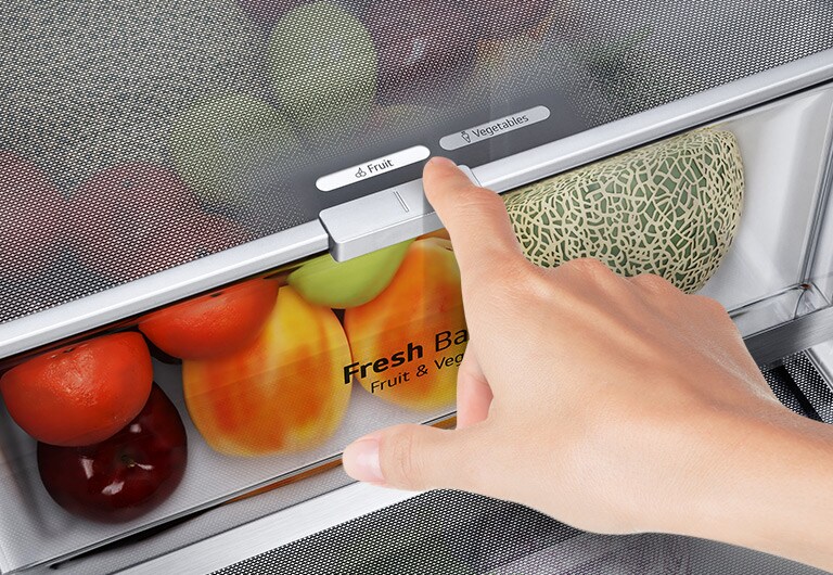 Hand adjusts freshness control in LG fridge drawer with tomatoes, apples, and melon, selecting fruit or vegetable setting.