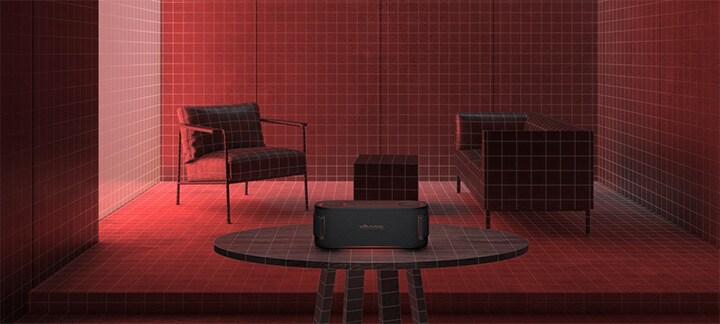 In a room filled with a red lighting a black sofa, a black ottoman  and a black armchair are placed behiind. In the fornt, xboom Bounce is placed on the top of black round table.