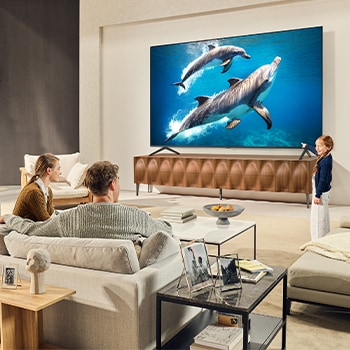A family sits on a couch facing a LG QNED TV sitting above a TV stand with a little girl pointing to a screen depicting two dolphins. The title talks about how you can enjoy high resolution on a massive screen.