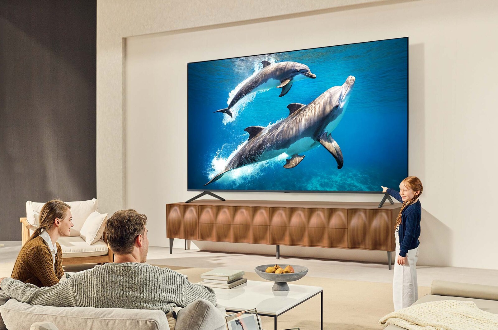 A family sits on a couch facing a LG QNED TV sitting above a TV stand with a little girl pointing to a screen depicting two dolphins. The title talks about how you can enjoy high resolution on a massive screen.