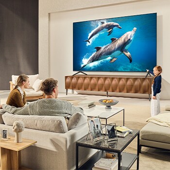 A family sits on a couch facing a LG QNED TV sitting above a TV stand with a little girl pointing to a screen depicting two dolphins. The title talks about how you can enjoy high resolution on a massive screen.