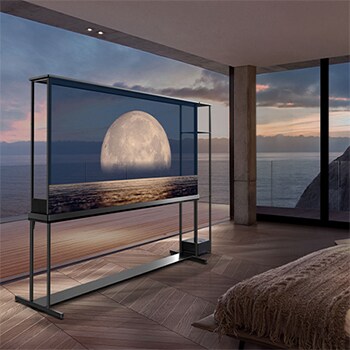 LG SIGNATURE OLED T displays a moon depicted above a shore on the transparent screen. The LG SIGNATURE OLED T sits in the middle of a room with a cliff side view that looks out to the sea. On the right is a bed and a lounge chair. 