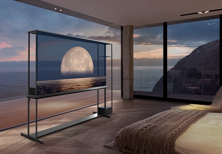 LG SIGNATURE OLED T displays a moon depicted above a shore on the transparent screen. The LG SIGNATURE OLED T sits in the middle of a room with a cliff side view that looks out to the sea. On the right is a bed and a lounge chair.