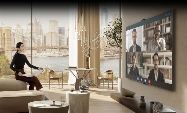 A woman is sitting on a luxurious sofa in a spacious living room. She is holding a laptop while facing a large screen on the opposite wall, indicating that she is in a video conference.