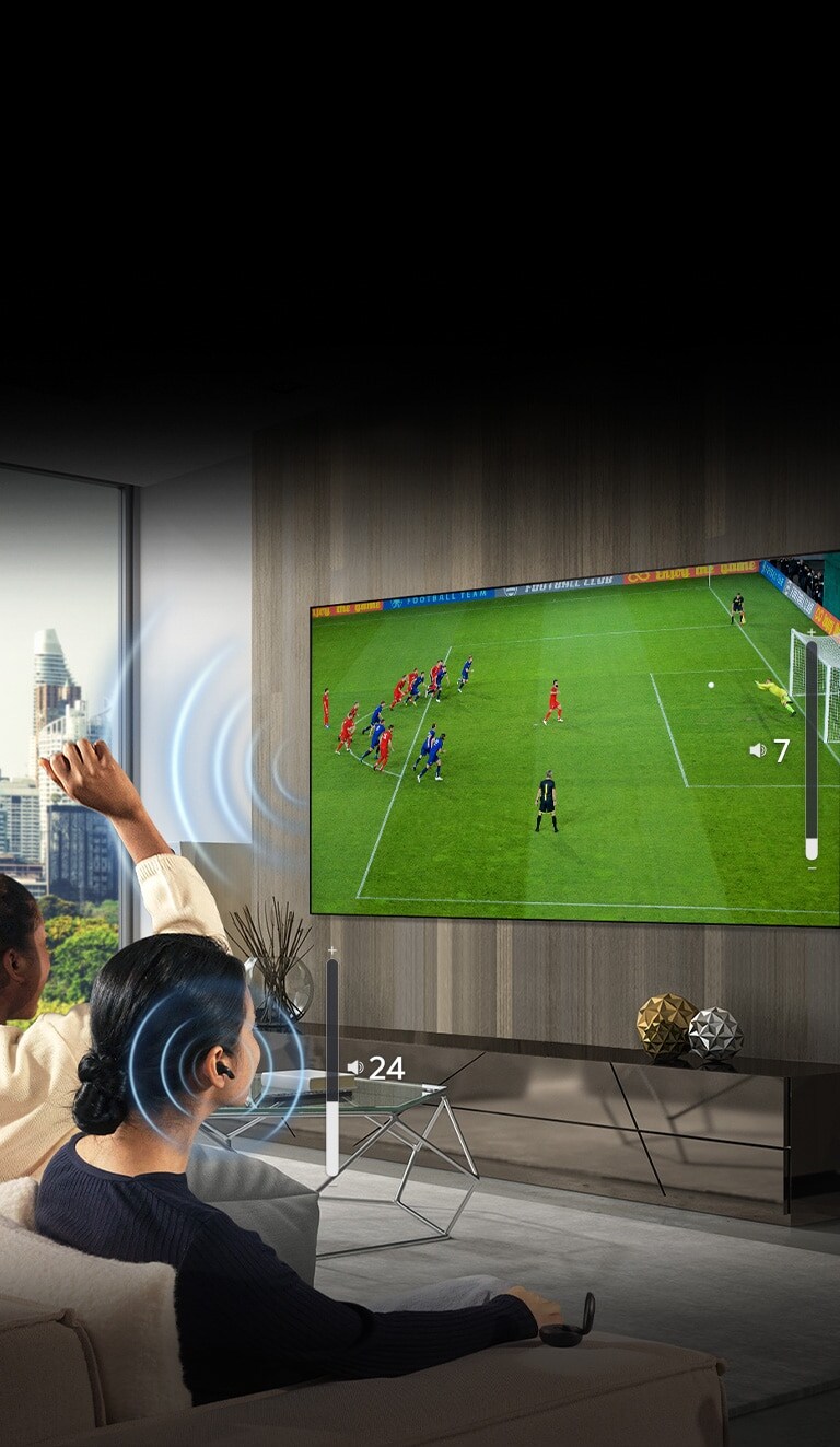 A group of people are sitting on a sofa watching a soccer game on TV. The woman on the far right is wearing earbuds and using them with a different volume than the TV, indicating that she is using both at the same time.