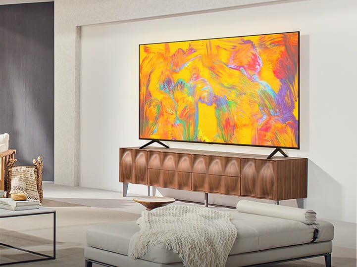 LG QNED TV berdiri di atas rak TV di ruang tamu yang nyaman. LG QNED TV menggambarkan latar belakang abstrak dalam warna oranye, kuning, ungu dan pink.