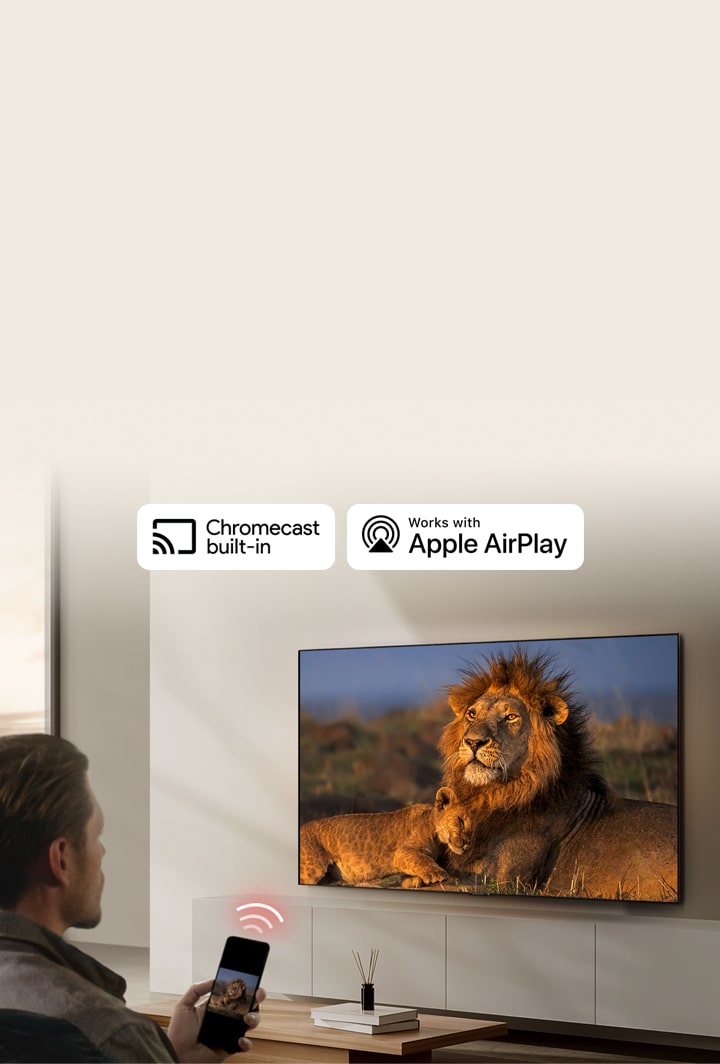 An LG TV mounted on a wall in a living room, displaying a lion and lion cub. A man sits in the foreground with a smartphone in his hand displaying the same image of lions. A graphic of three neon red curved bars is displayed just above the smartphone pointing toward the TV.