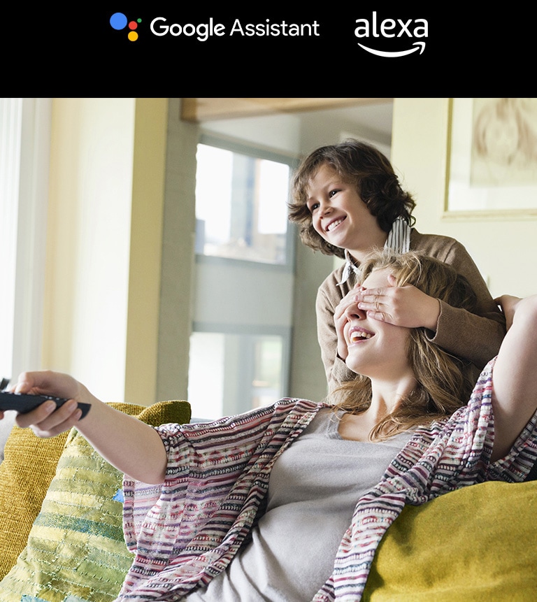 A kid is covering his mother’s eyes who is holding a TV remote. There is Google Assistant logo and alexa logo above image.