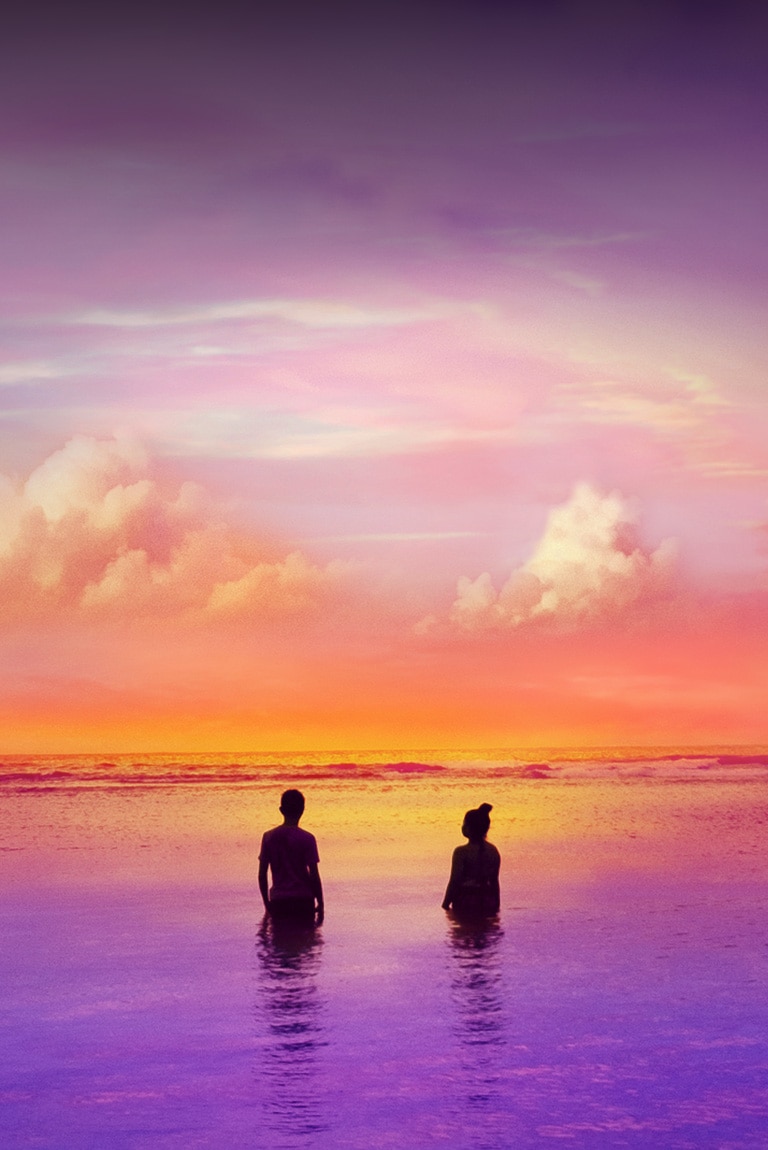 Two people are standing inside ocean – a slightly purple ocean. It’s a sunset time.