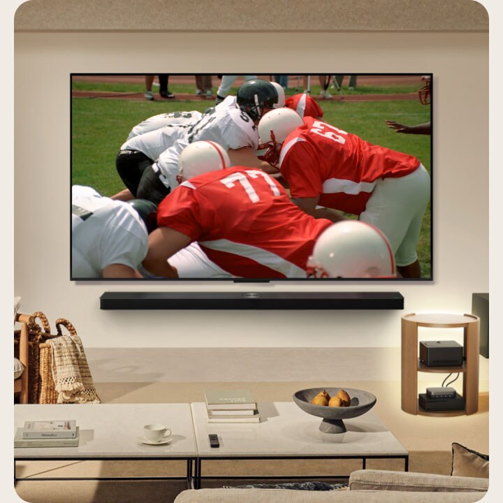 Salotto con il TV LG True Wireless montato sulla parete. Sullo schermo viene trasmessa una partita di football americano. Inoltre, la configurazione del televisore include un LG Soundbar montato a parete. Quasi nascoste alla vista, le altre periferiche sono collegate a uno Zero Connect Box posizionato su un tavolino. L’intero salotto è pulito, ordinato e privo di cavi. 