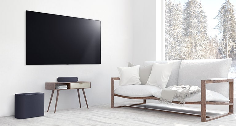 Una soundbar è collocata su un piccolo tavolino, mentre un subwoofer si trova sulla sinistra e un TV sopra la soundbar. L’intera stanza è bianca.