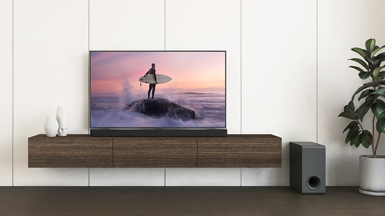 Un TV LG e una soundbar LG sono posizionati su un ripiano marrone, mentre il subwoofer è sul pavimento. Lo schermo del TV mostra un surfista in piedi su uno scoglio.