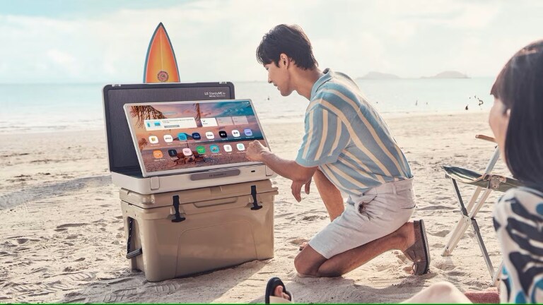 Un uomo che installa un dispositivo portatile LG su un frigorifero da spiaggia, con una tavola da surf sullo sfondo. Il dispositivo mostra un'interfaccia utente con varie app.