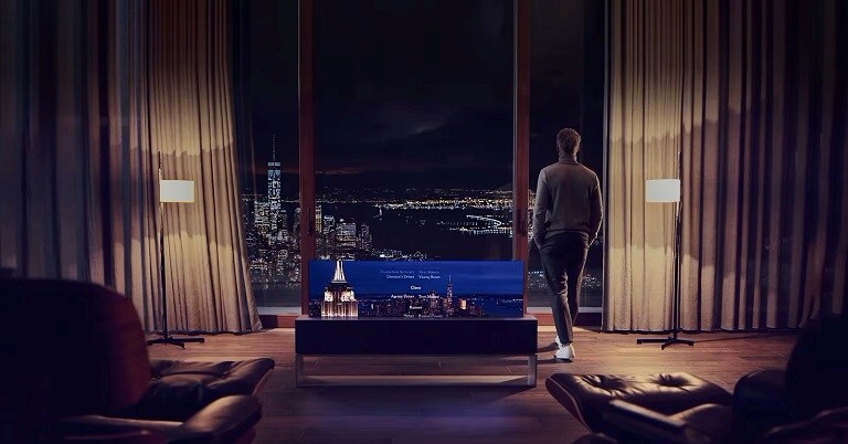 Un uomo in piedi davanti a una grande finestra in un elegante salotto con vista sulla città, mentre una TV LG con schermo arrotolabile mostra i titoli di coda di un film.