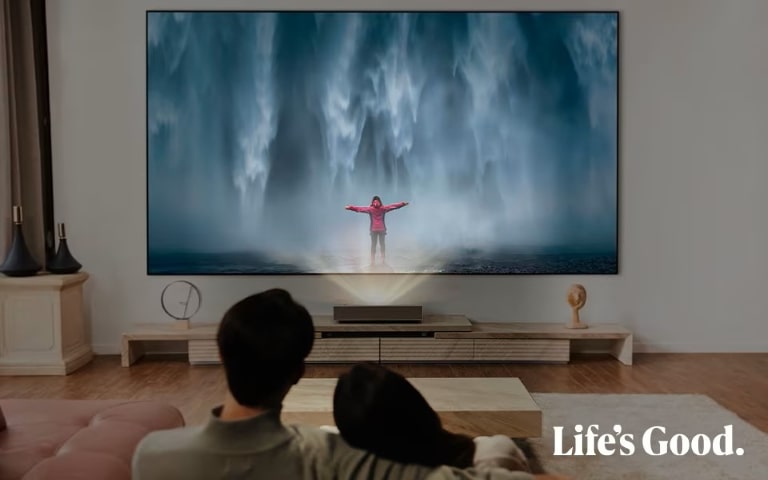 Una coppia seduta su un divano in una sala cinema domestica, che guarda una TV a grande schermo che mostra una persona in giacca rossa di fronte a una cascata. Il logo LG e lo slogan "Life's Good" sono visibili nell'angolo.