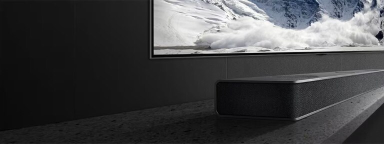 Una elegante soundbar posizionata su una superficie in pietra scura sotto una grande TV a parete che mostra una scena di montagna innevata.