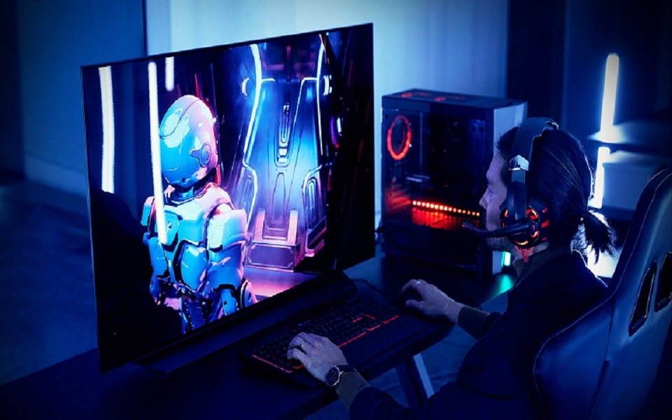 Un gamer con un headset gioca a un gioco di fantascienza su un grande schermo, con un PC luminoso accanto a lui in una stanza poco illuminata.