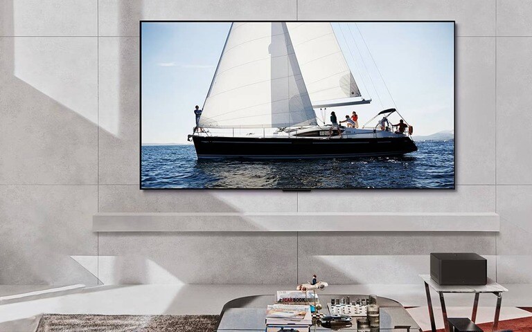 Un TV LG montato su una parete elegante del soggiorno, che mostra una vivida scena di una barca a vela sull'oceano, con un arredamento moderno e minimalista, inclusa una mensola bianca essenziale e un tavolino con riviste e oggetti decorativi.