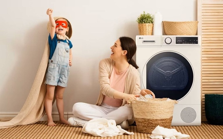 Una madre felice che piega il bucato accanto a un bambino vestito da supereroe, vicino a un'asciugatrice LG a pompa di calore, creando un'atmosfera giocosa e accogliente in lavanderia.