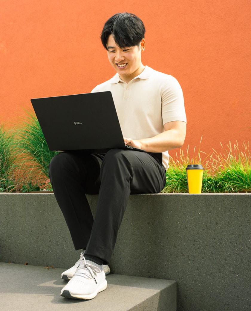 Un uomo seduto all'aperto contro un muro arancione brillante, lavora al suo laptop LG gram con una tazza di caffè accanto, godendosi la giornata soleggiata.