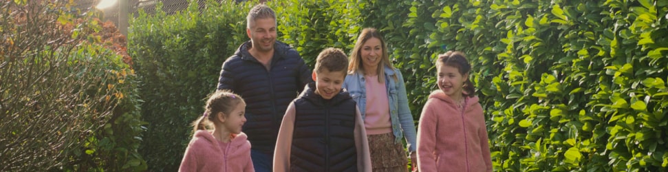 Famiglia di cinque persone che si gode una passeggiata all'aperto, sorridente e in abbigliamento casual, circondata da siepi verdi.