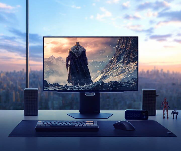 ハイエンドゲーミングモニターに、雪山を背にしたフード付きの戦士が立つ、壮大なファンタジーゲームのワンシーンが映し出されている。モニターは、高層マンションのスタイリッシュなデスクに設置されており、背景には夕暮れの都市のスカイラインが広がっている。モニターの周囲にはスピーカー、キーボード、ヘッドセット、時計、小さなアクションフィギュアが配置され、没入感とスタイルを兼ね備えたゲーミング環境を演出している。