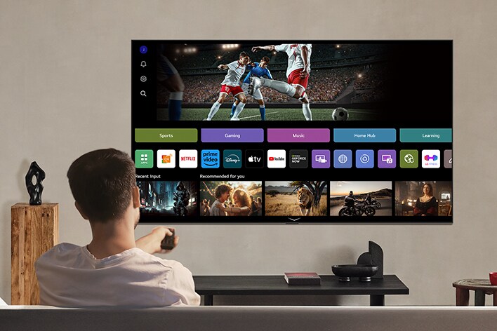 Человек на диване протягивает пульт дистанционного управления. Телевизор LG AI TV на стене с webOS от LG на экране.