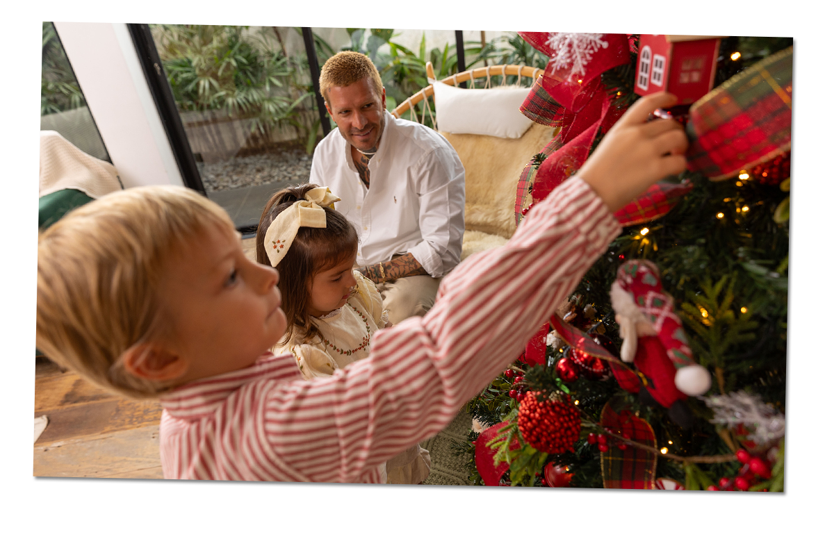 Un padre y sus hijos se divierten decorando un árbol de Navidad. El niño sostiene un adorno para colocarlo en el árbol.