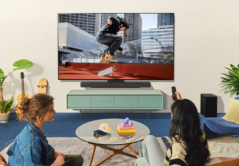 Sala de estar con dos personas viendo a un patinador en la LG OLED TV montada en la pared, rodeada de plantas, libros y decoración moderna