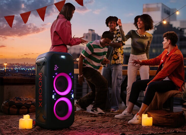 Grupo de amigos reunidos alrededor de una bocina LG XBOOM en una fiesta en la azotea, con anillos de luz rosa iluminando la bocina.