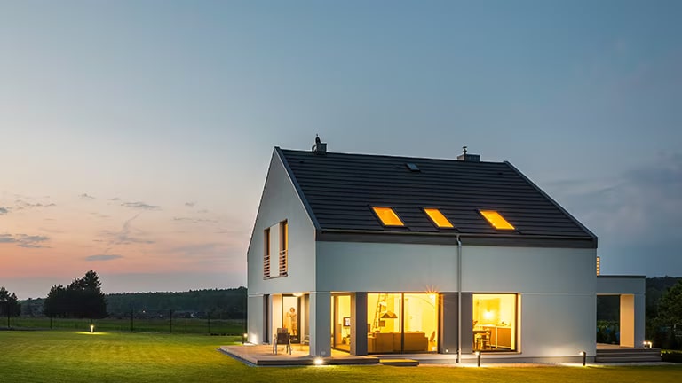 En medio de una exuberante zona de hierba, una gran casa blanca con un techo negro emana luz, iluminando brillantemente el paisaje circundante.