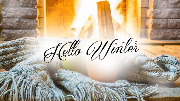 Chimenea encendida con una manta tejida en primer plano y el texto 'Hello Winter' superpuesto, creando una atmósfera cálida y acogedora.
