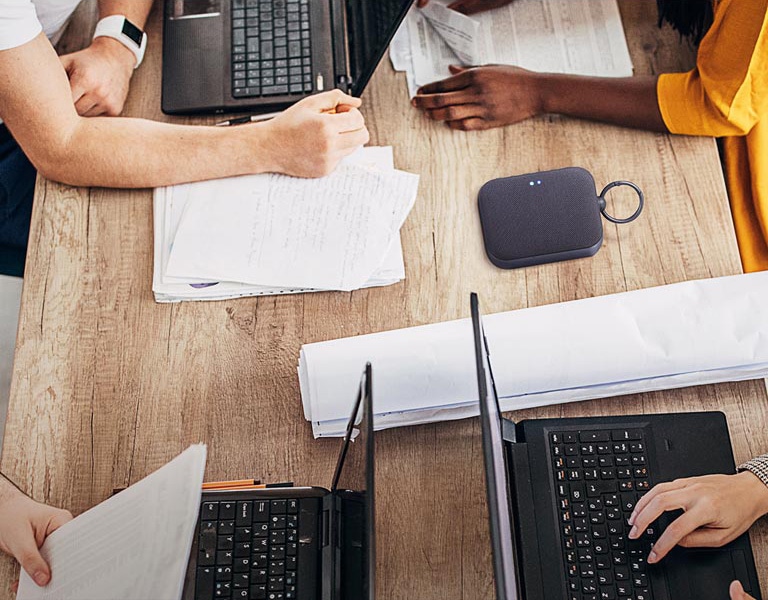 4 personas están trabajando en un escritorio, cada uno con su propia computadora portátil. Los rostros no se muestran, algunos están tecleado, algunos están escribiendo. El altavoz XBOOM Go PM1 se coloca sobre un escritorio.