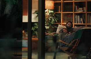 Una escena donde la cámara captura a un hombre y una mujer acostados juntos en el sofá de la sala de estar desde una distancia.