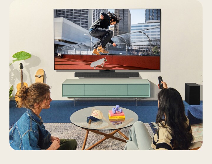 Dos mujeres sentadas en un sofá frente a una LG TV montada en la pared de una sala colorida, y una de ellas apunta con el control remoto a la pantalla de la televisión. 