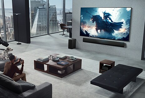 Woonkamer met iemand die een videogame speelt op zijn LG True Wireless TV met LG Soundbar die op de wand zijn gemonteerd. Hij heeft een controller in zijn hand, maar de spelcomputer is niet meteen zichtbaar. Er zijn ook geen draden zichtbaar. De Zero Connect Box is opgeborgen en nauwelijks zichtbaar onder een bijzettafeltje.
