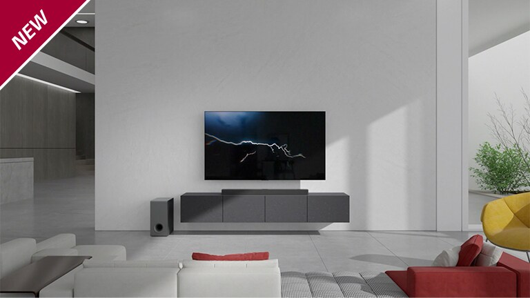 De soundbar is geplaatst op een grijze kast met een tv die in de woonkamer aan de muur hangt. Een draadloze subwoofer is links op de vloer geplaatst en het zonlicht komt van rechts in beeld. Een wit met rood gekleurde lange bank is tegenover de tv en soundbar geplaatst. In de linkerbovenhoek staat NEW mark.