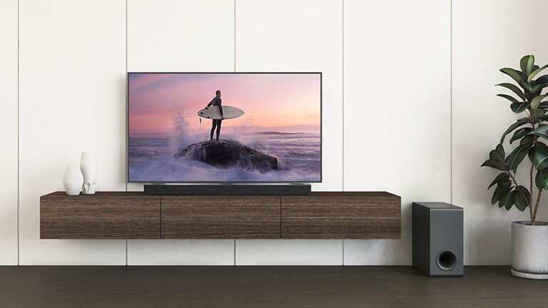 Een LG TV en LG soundbar staan op een bruine plank en de subwoofer staat op de vloer. Het scherm toont een surfer die op de rots staat.