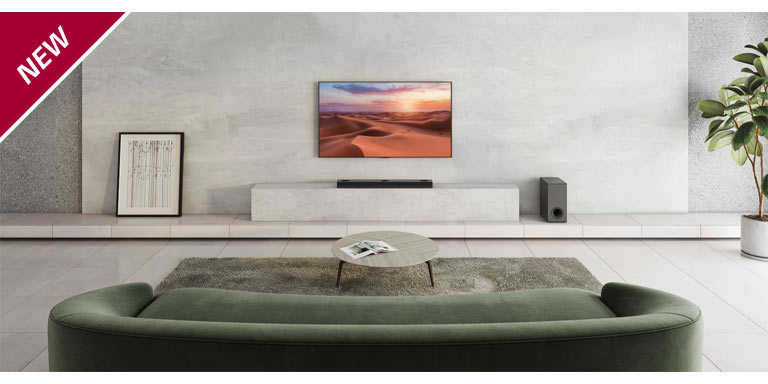 De tv is opgehangen aan de witte pilaar in het midden van de woonkamer. Hieronder staat de LG Soundbar op de witte plank. Op de vloer rechts onder de soundbar staat een draadloze subwoofer. Achter de groene bank zijn links en rechts op de salontafel twee achterspeakers geplaatst. In de linkerbovenhoek staat NEW mark.