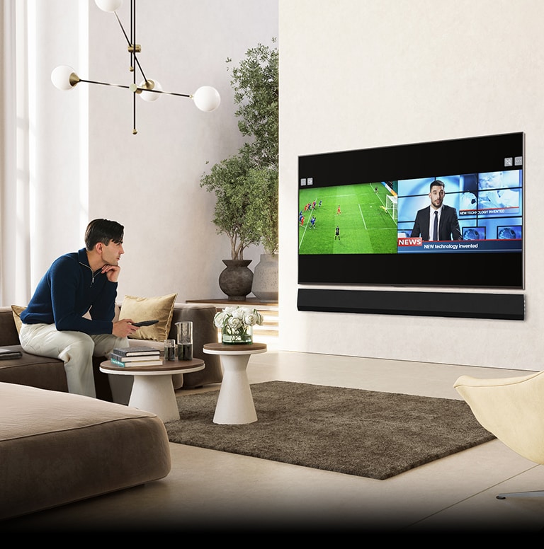 Een man zit op een luxe en ruime huiskamerbank en kijkt naar de tv. Het tv-scherm toont een split-screen weergave van een voetbalwedstrijd en een nieuwsprogramma.