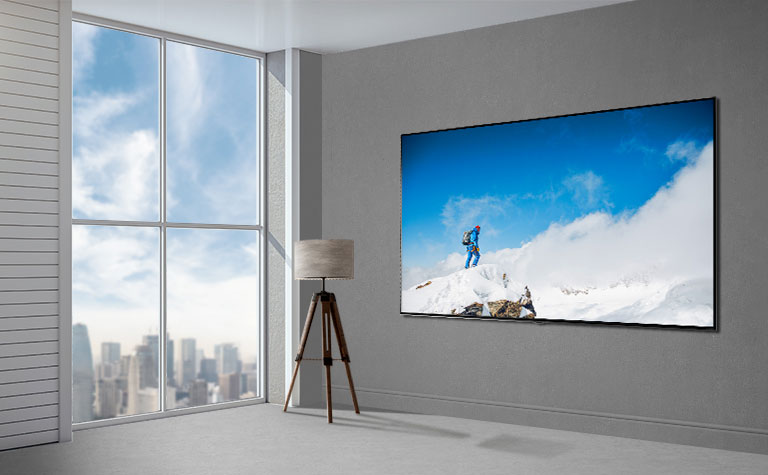 Een flatscreen-tv aan een grijze muur naast een raam van vloer tot plafond en een stoel. Links van de tv staat een lamp en op het scherm is een wandelaar te zien op een met sneeuw bedekte rots staat.