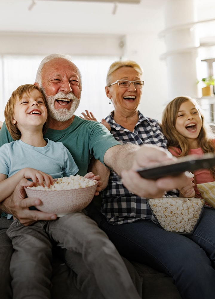 Una familia sentada riendo mientras el abuelo apunta con el control remoto.