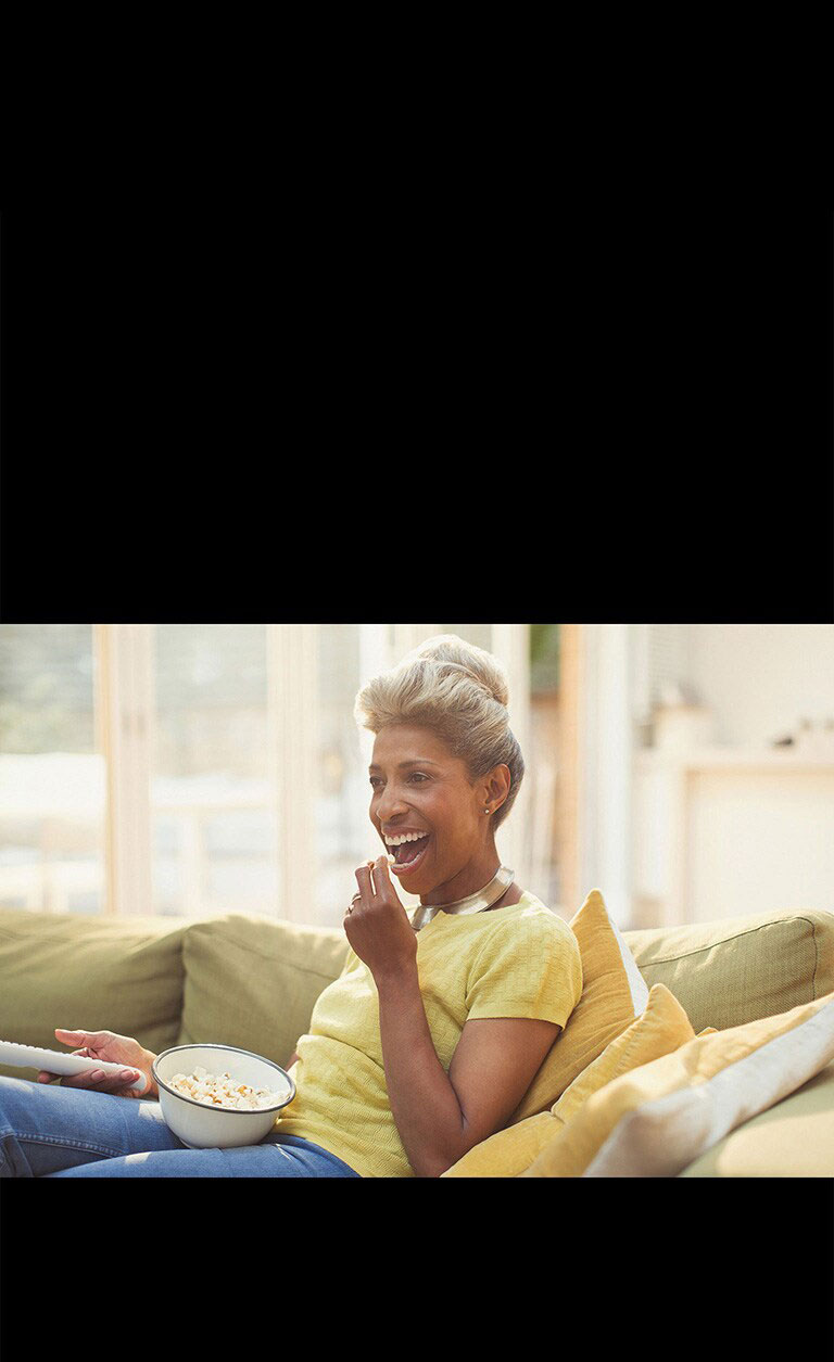 Una mujer está viendo la televisión, y sostiene un control remoto. También está comiendo palomitas de maíz.