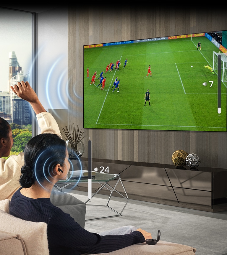 Un grupo de personas está sentada en un sofá viendo un partido de fútbol en la televisión. La mujer en el extremo derecho usa auriculares y los usa con un volumen diferente al del televisor, lo que indica que estan usando ambos al mismo tiempo.