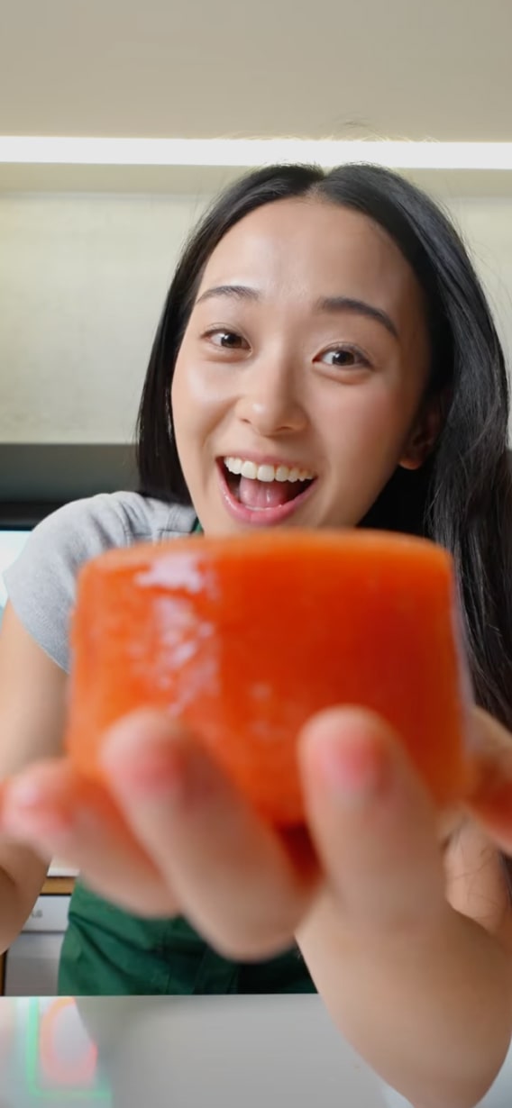 Mujer con cabello largo y oscuro sonriendo y sosteniendo un trozo de sushi cerca de la cámara.