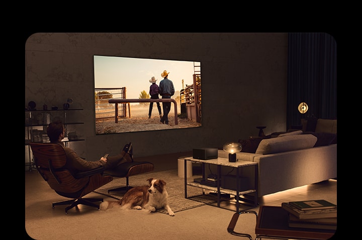 Un hombre viendo una película de vaqueros con su perro en un acogedor espacio habitable durante la noche. No hay cables alrededor de la LG OLED TV montada en la pared y la Caja de Conexión Cero se ubica perfectamente sobre una mesa auxiliar. Y luego, la imagen pasa a otra imagen que muestra a un hombre viendo una película de vaqueros con la  LG OLED TV sobre un soporte de piso en un espacio acogedor y oscuro. No hay cables alrededor de la LG OLED TV y su soporte de piso, y la Caja de Conexión Cero se ubica ordenadamente sobre una mesa auxiliar.