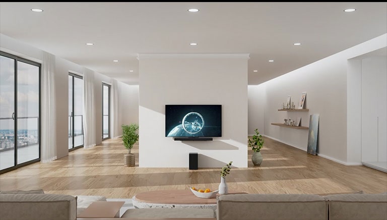 La barra de sonido y el televisor están en el pilar blanco en el centro de la sala de estar. En el piso de madera, justo debajo de la barra de sonido, hay un woofer negro. En el lado izquierdo hay ventanas de piso a techo con excelente vista y cortinas blancas. En el lado derecho hay estantes de madera con adornos y una imagen en el suelo.