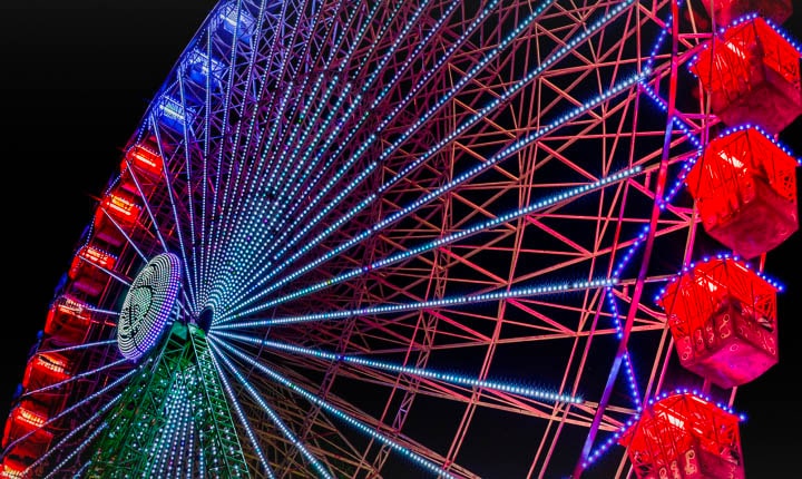 Una noria ligeramente descolorida se ilumina durante la noche, con destellos de colores que comienzan en el lado izquierdo de la pantalla y se mueven a través de ella. A medida que pasan los destellos de colores, la pantalla pasa de colores nublados a colores brillantes y vivos.
